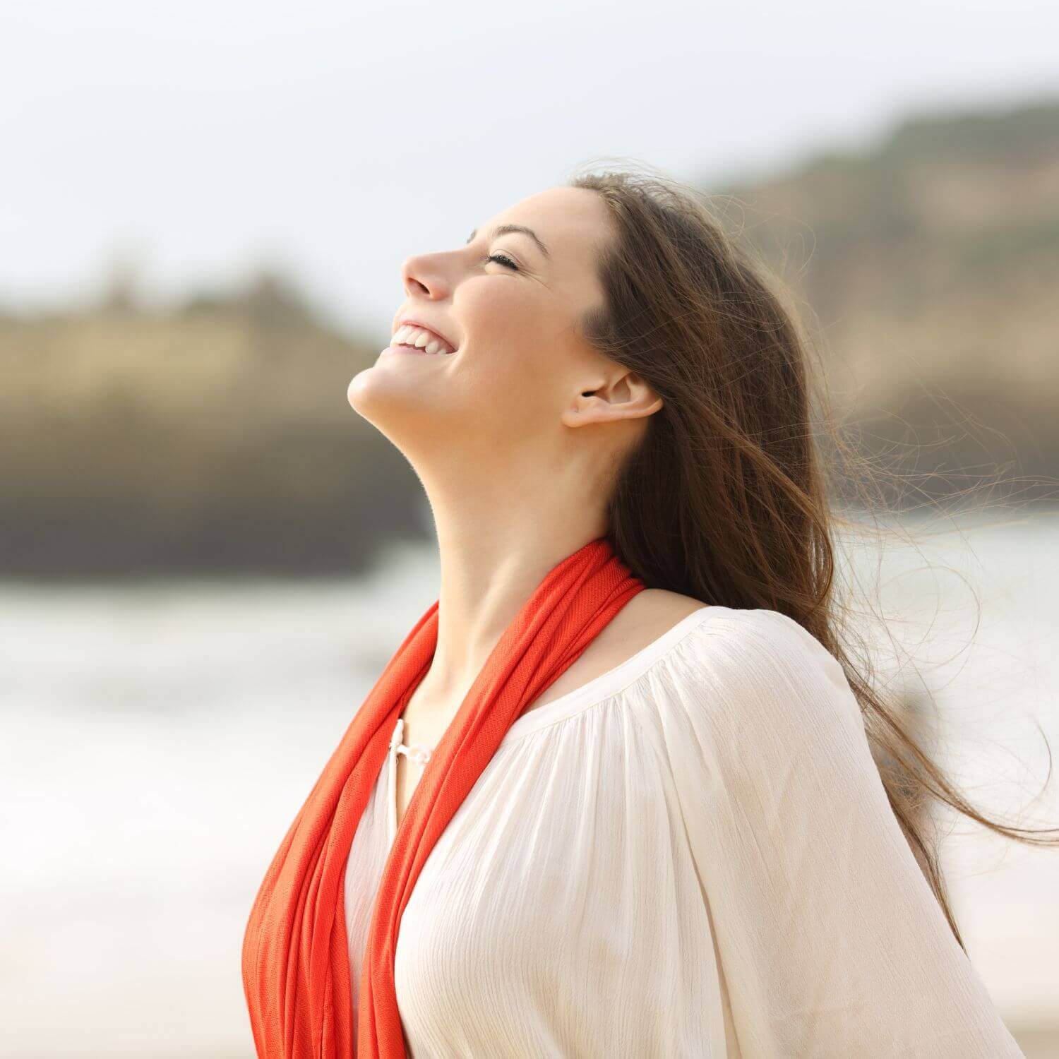 Mulher sorridente na praia usando um lenço vermelho, parecendo relaxada e feliz.