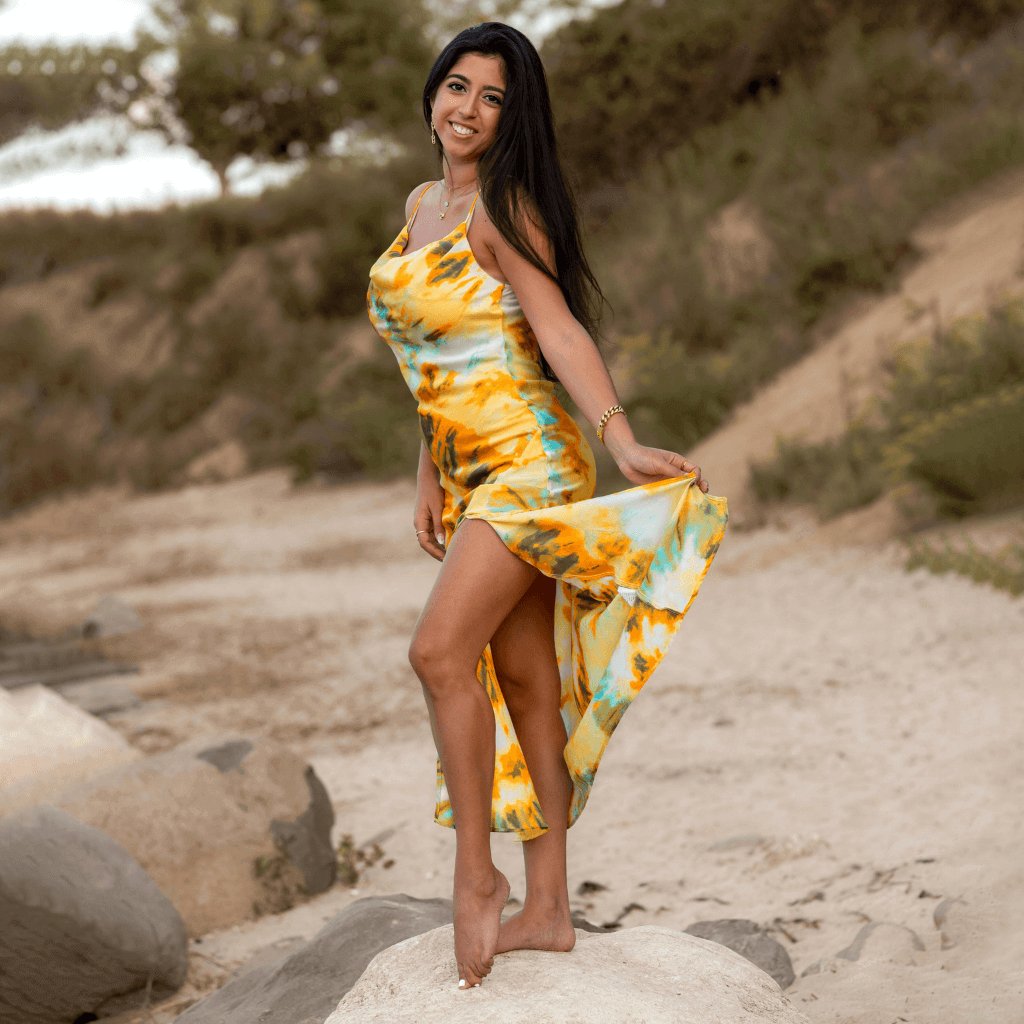 Mulher em vestido colorido amarelo e azul, posando na praia com olhar alegre e natural.